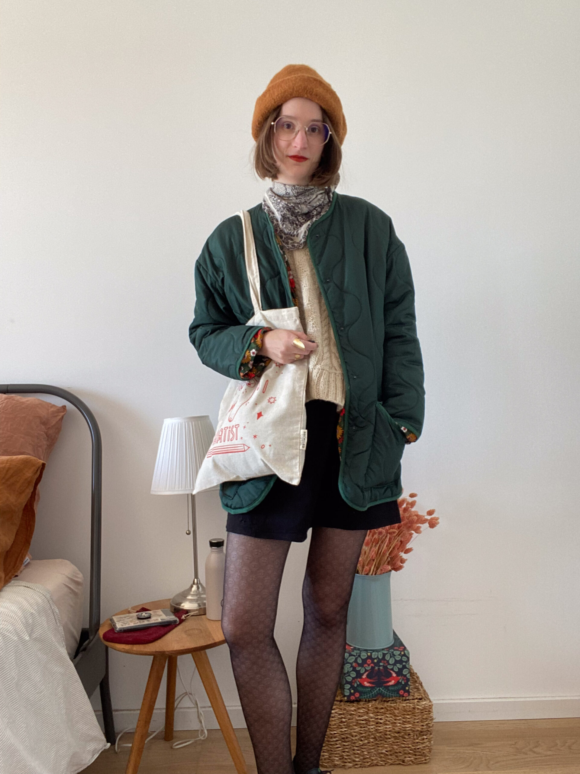 Photographie d'une femme portant une robe noire, un pull à torsade sans manche écru, une veste matelassée vert sapin, un chapeau marron et une totebag